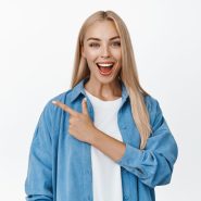 Amazed blond girl points finger left and looks impressed, shows advertisement, stands against white background.